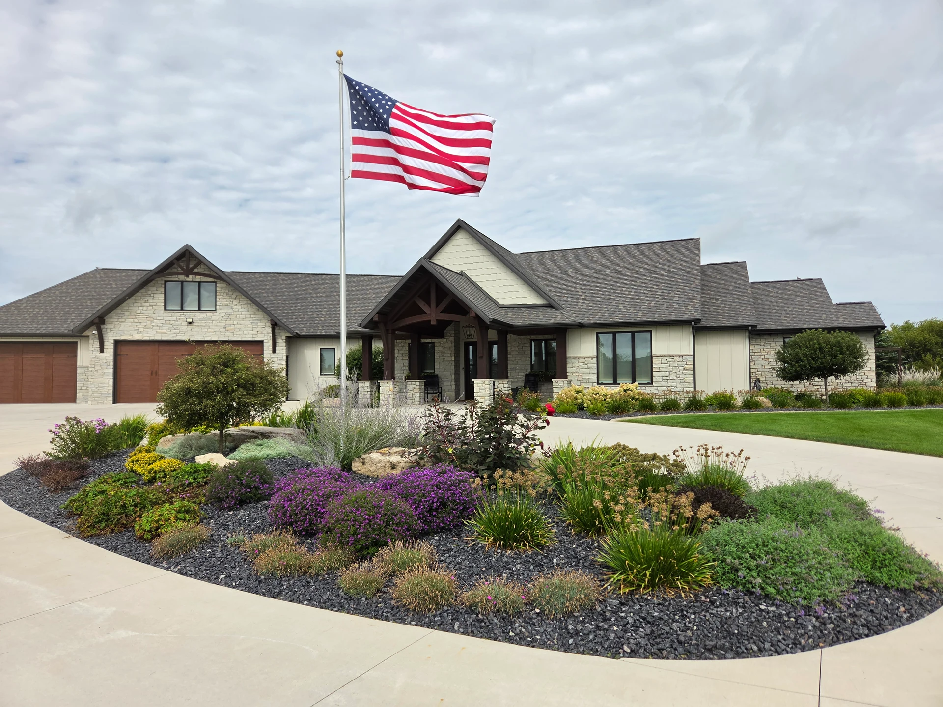 Colorful perennial flowers, shrubs, boulders, and decorative stone surrounding an American flagpole in a curved driveway island in front of a stone-sided home.