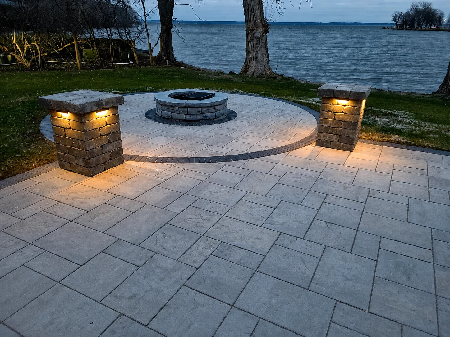 Stone firepit at dusk with illuminated stone pillars beside a lake