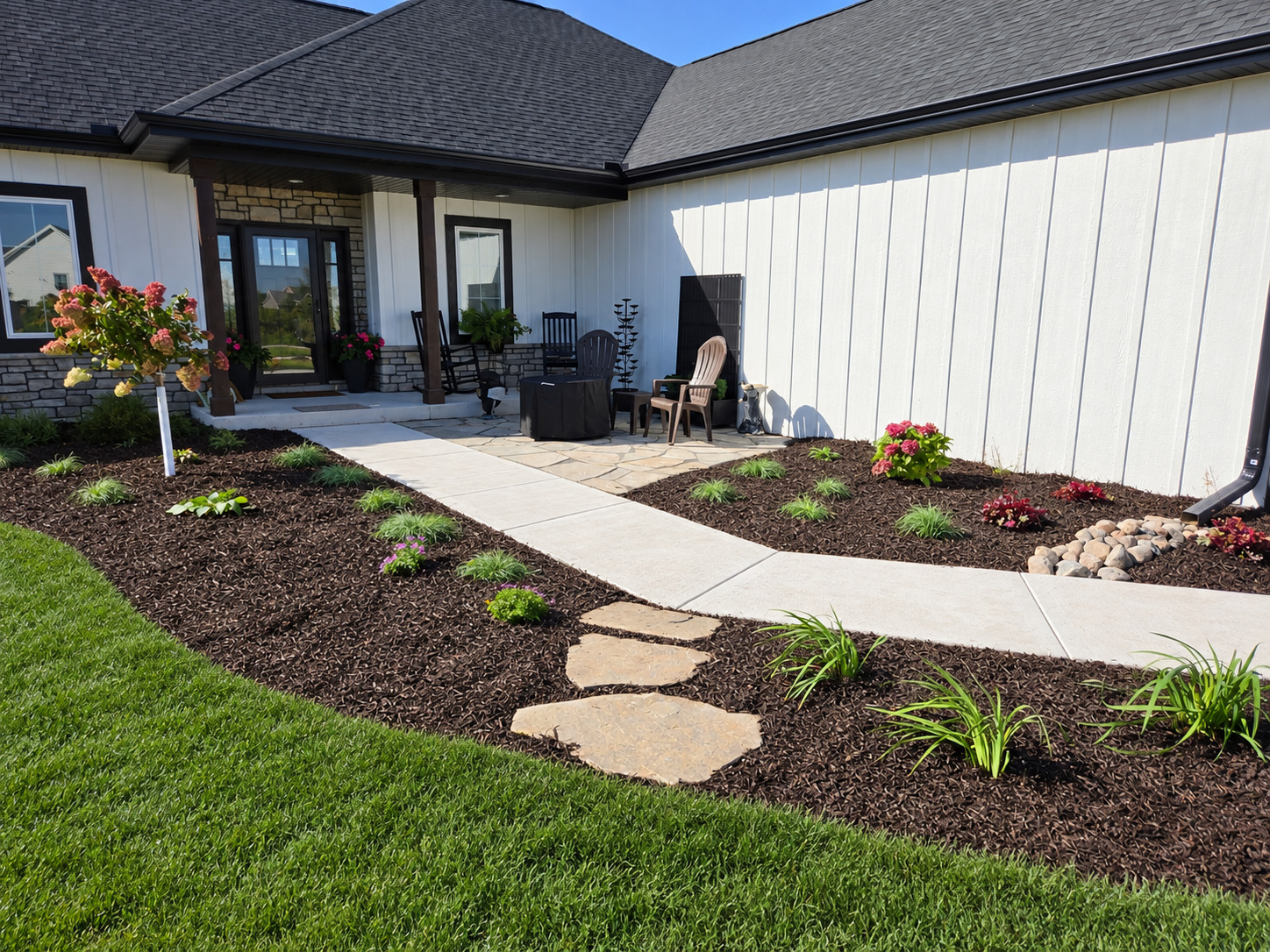 Curved concrete walk through mulched planting beds with flowering plants, ornamental grasses, and a small tree at the front entry of a white home.