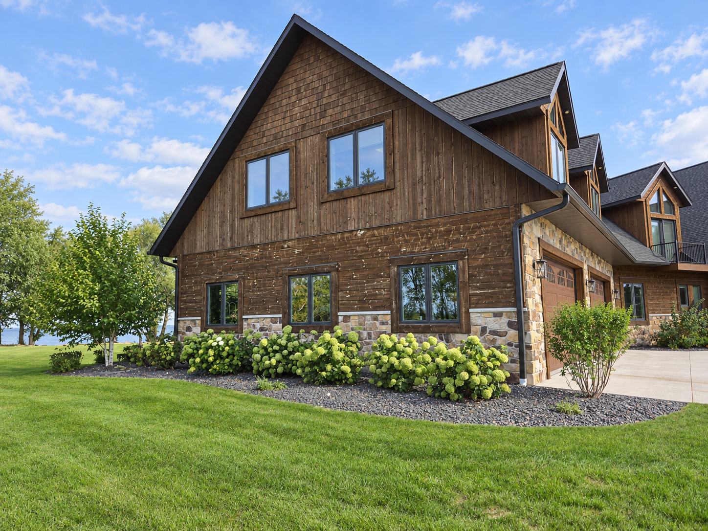 Flowering hydrangeas in a curved decorative-stone bed beside a wood-and-stone lake home, bordered by green lawn.