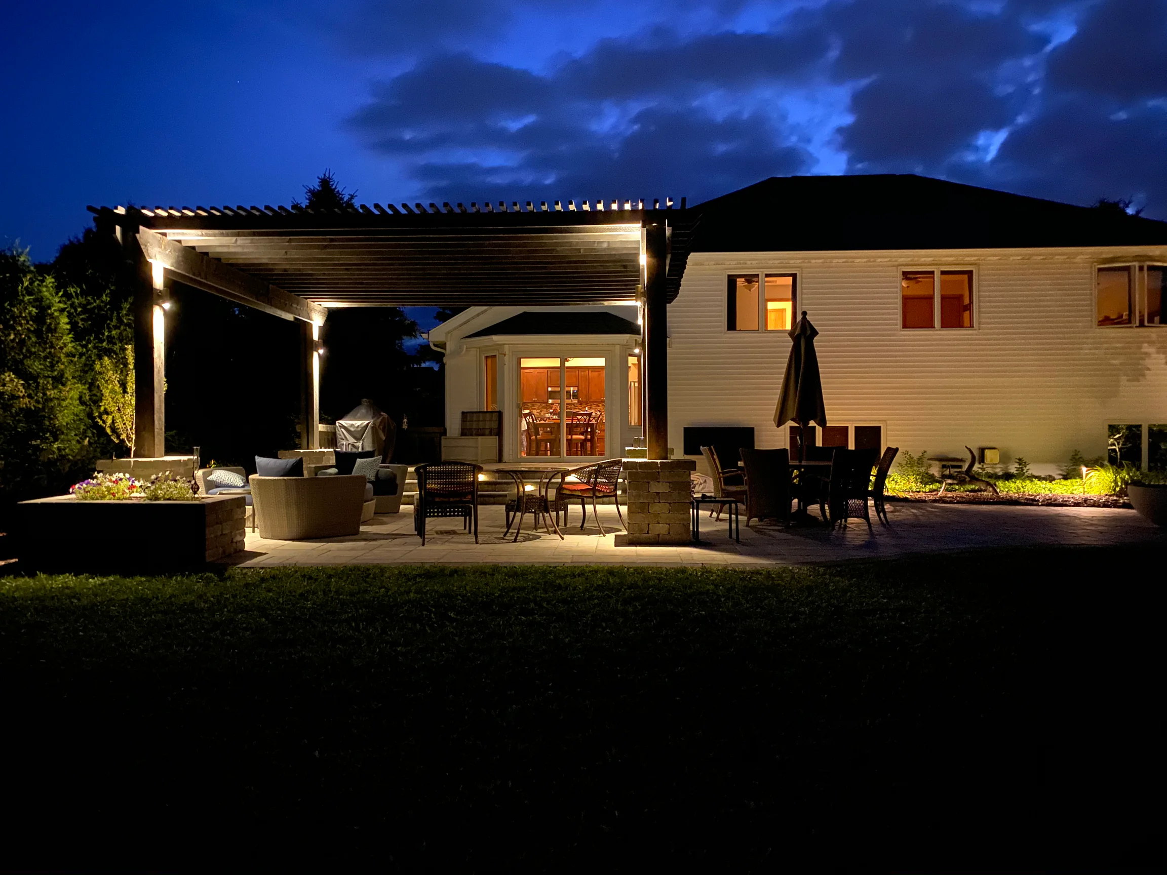 Wide paver patio and pergola illuminated with warm landscape lighting beneath a deep blue evening sky