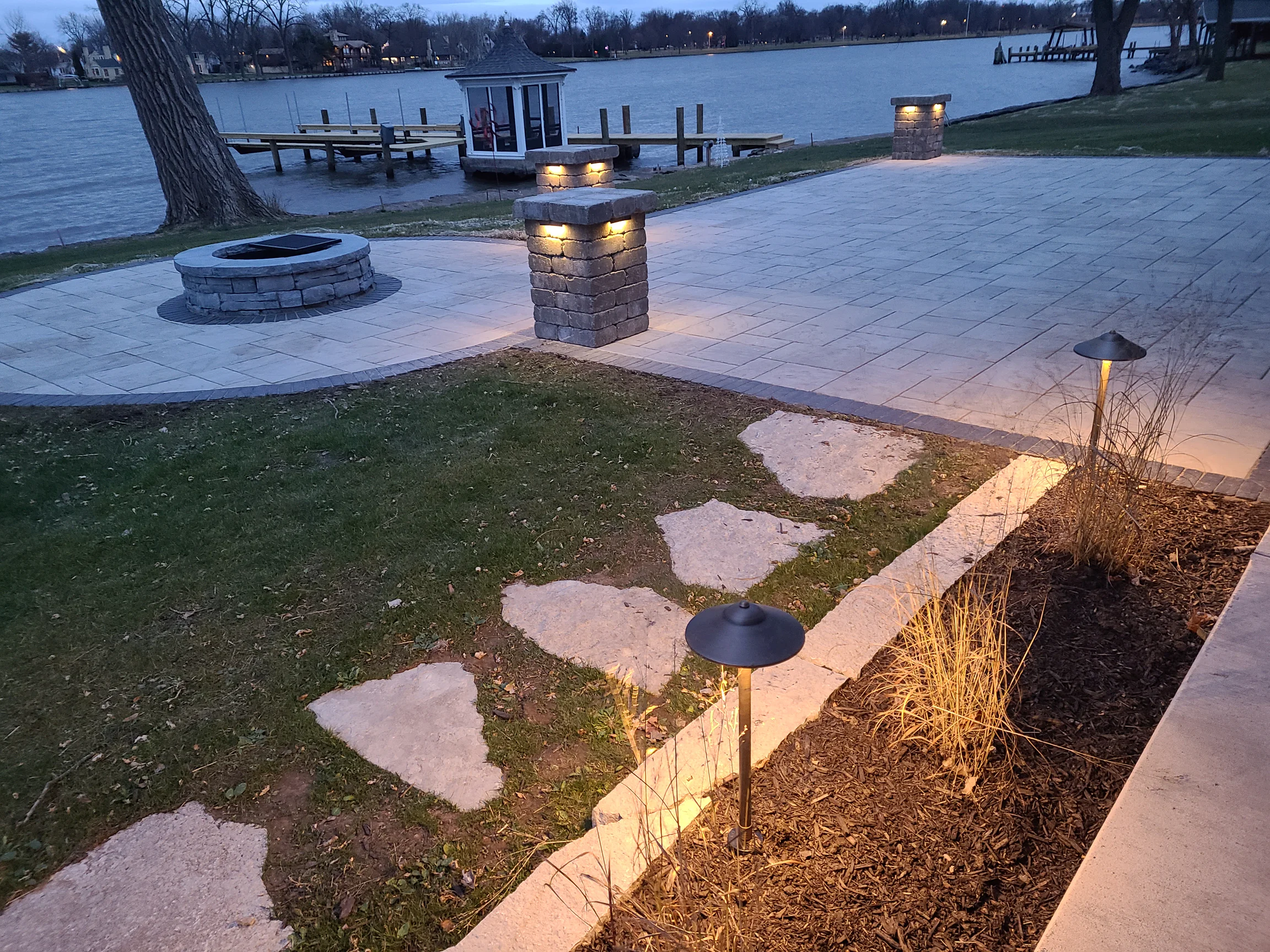 Lakeside paver patio and firepit with illuminated stone pillars and warm path lights at dusk