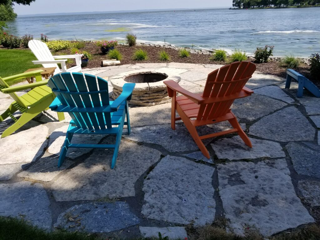 Colorful Adirondack chairs surround a stone fire pit beside the lake.
