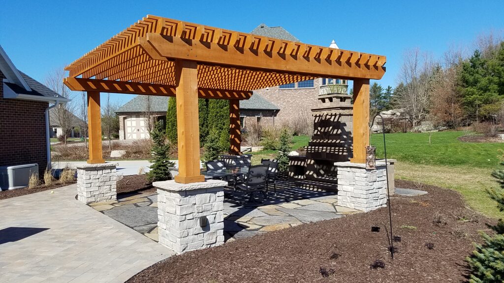 Wide cedar pergola with stone piers beside a masonry outdoor fireplace.