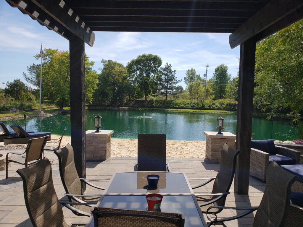 Dark-stained pergola shelters outdoor seating beside a landscaped pond.