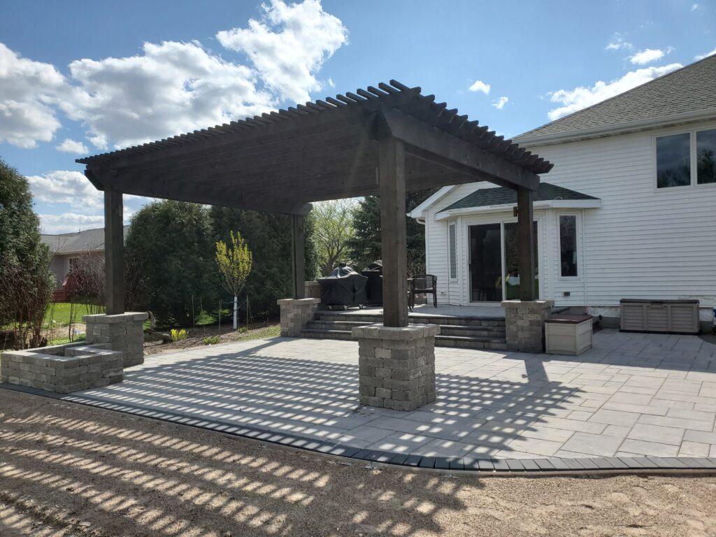 Dark pergola with stone columns covers a spacious paver terrace.