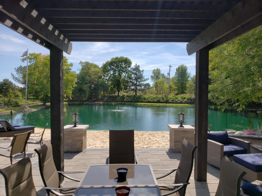 Covered paver patio under a dark pergola overlooking the pool.